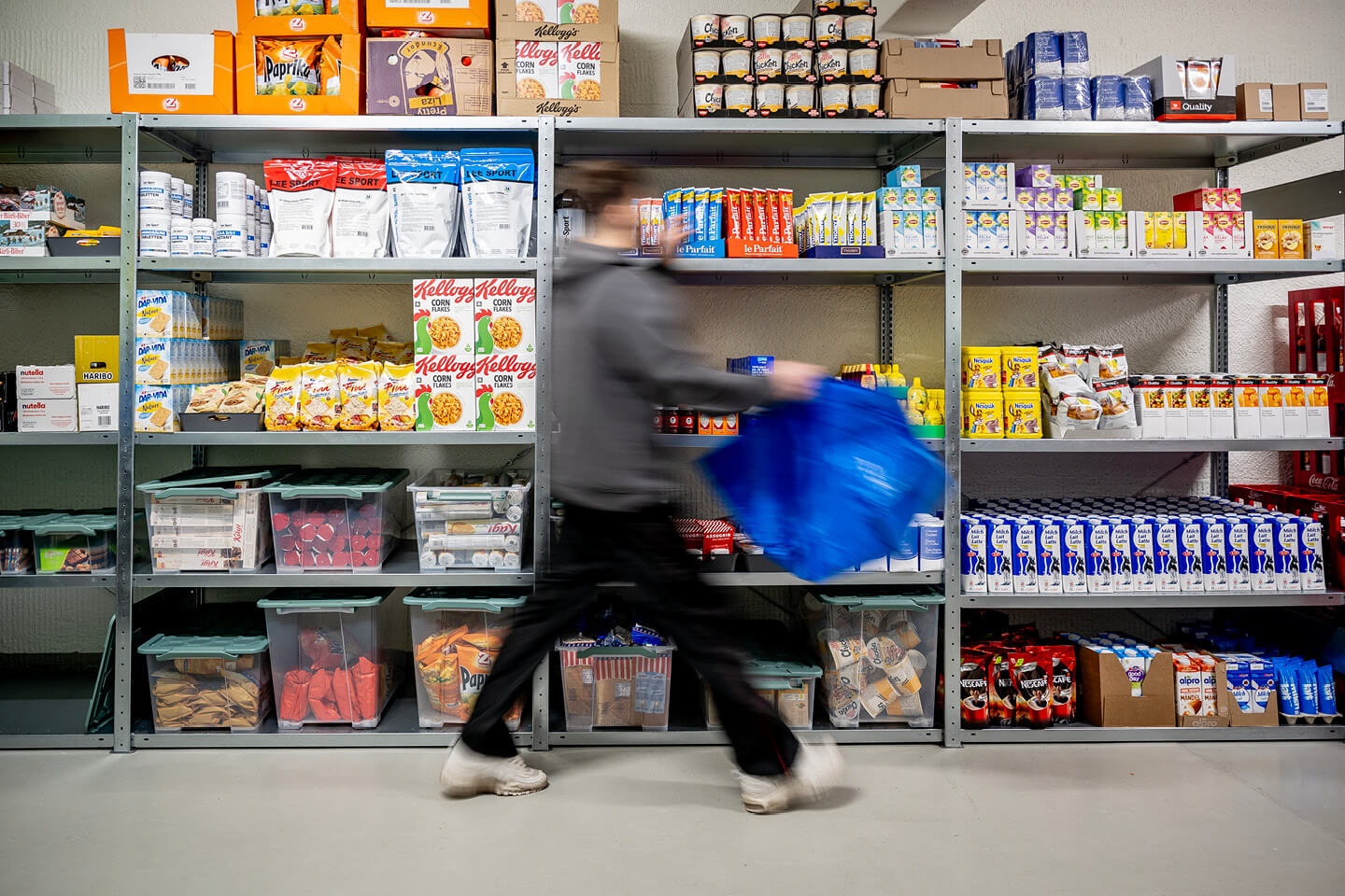 Inhaftierter im Gefängnisshop. Die Regale sind mit diversen Lebensmitteln und Artikeln zur Körperpflege gefüllt. Die Person trägt eine grosse Tasche als Einkaufskorb mit sich.